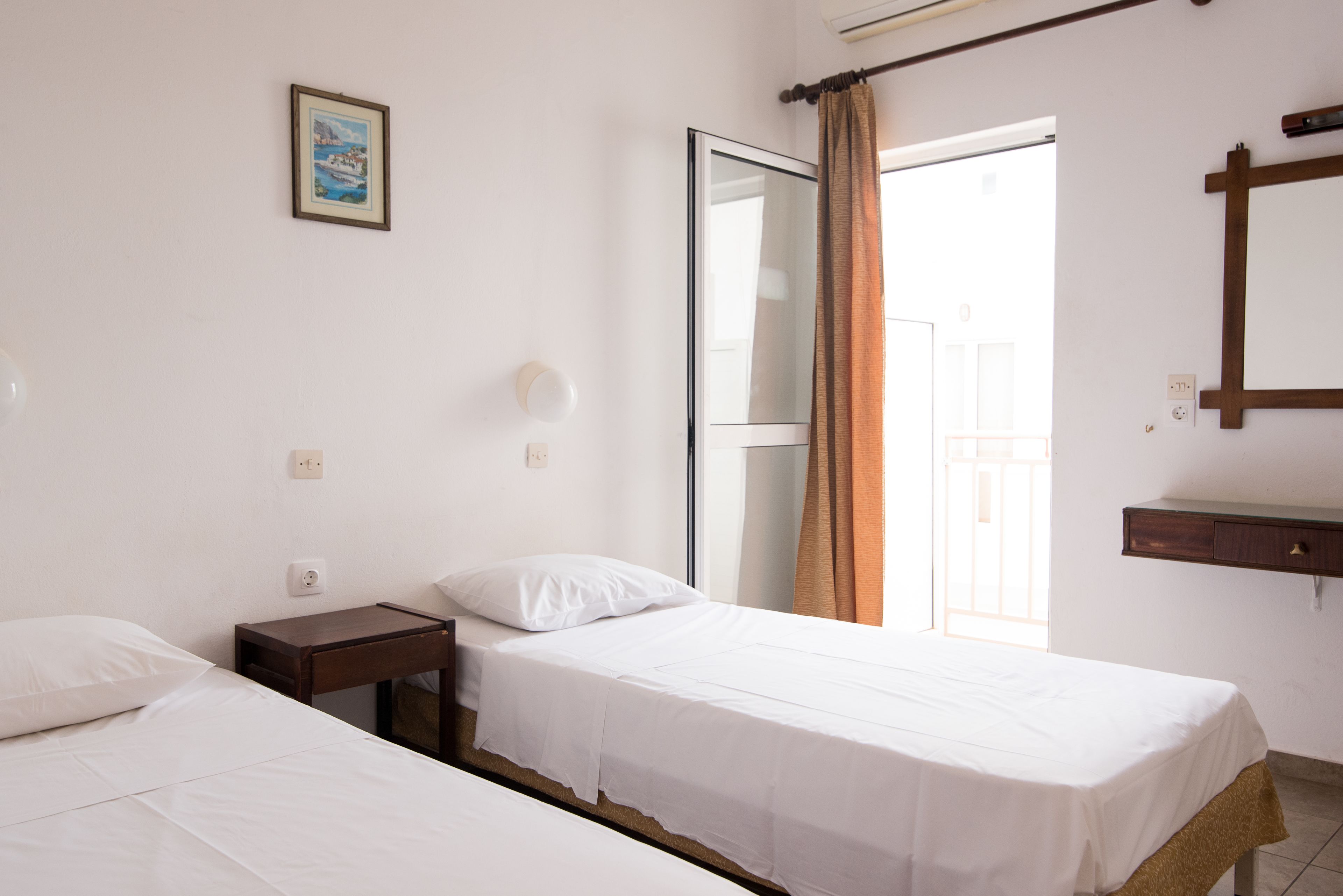 Habitación de hotel con dos camas individuales, una mesita de noche, una lámpara de pared, un cuadro y una puerta corredera a un balcón.