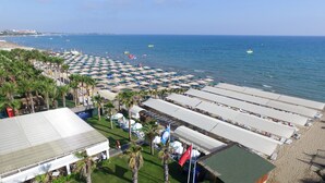 A beachside hotel with rows of beach umbrellas and lounge chairs, a pool area, and a view of the sea.