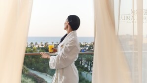 A person in a white robe standing by a window with a view of the sea and a cityscape.