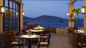 An outdoor dining area with tables and chairs, overlooking a body of water with boats and a distant island.