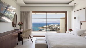 Quarto de hotel moderno com uma cama grande, uma escrivaninha e uma cadeira. Vê-se o mar e edifícios pela janela.
