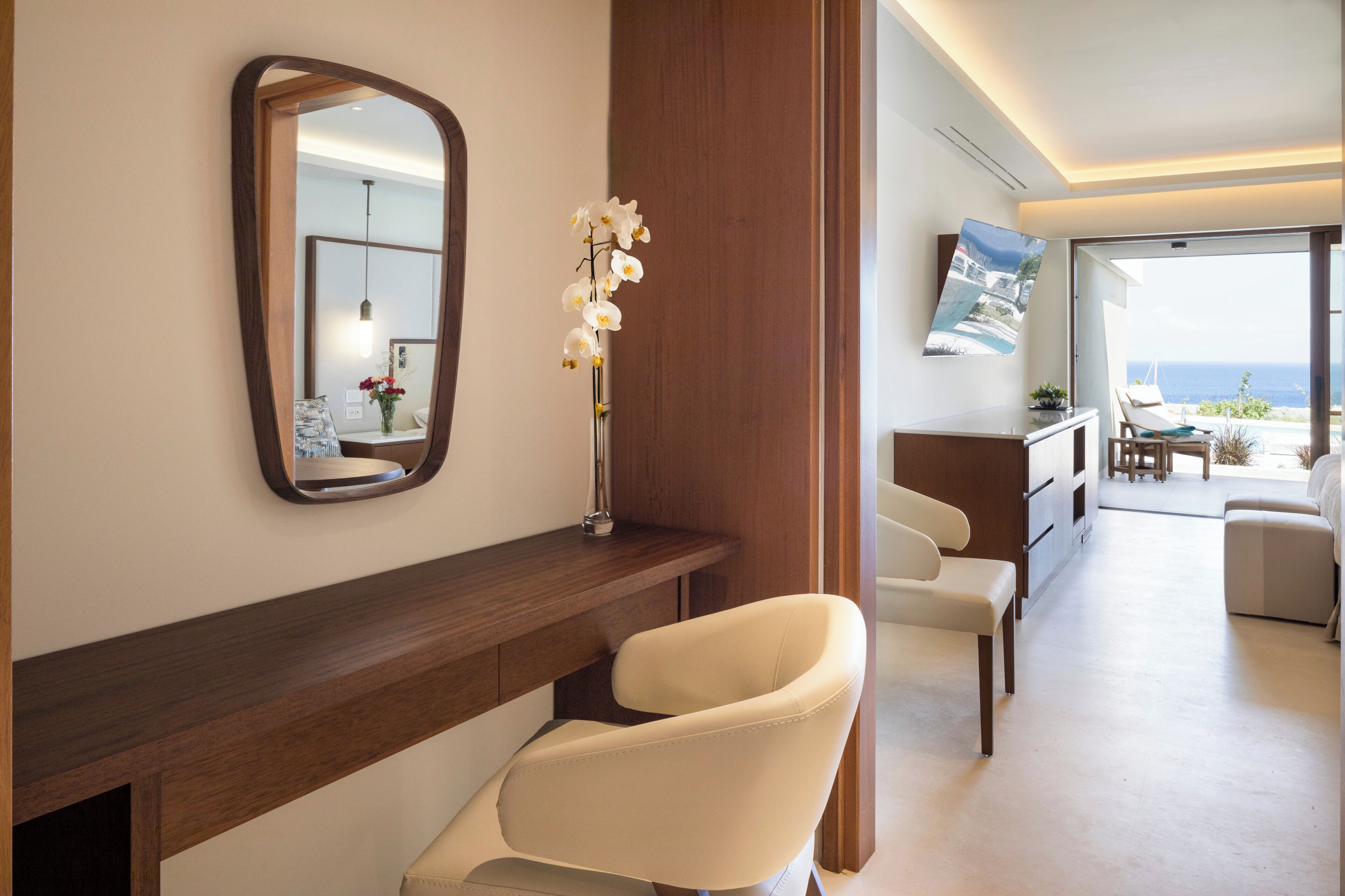 Une chambre d’hôtel moderne avec une console en bois, une chaise blanche, un miroir et un vase contenant des fleurs blanches.