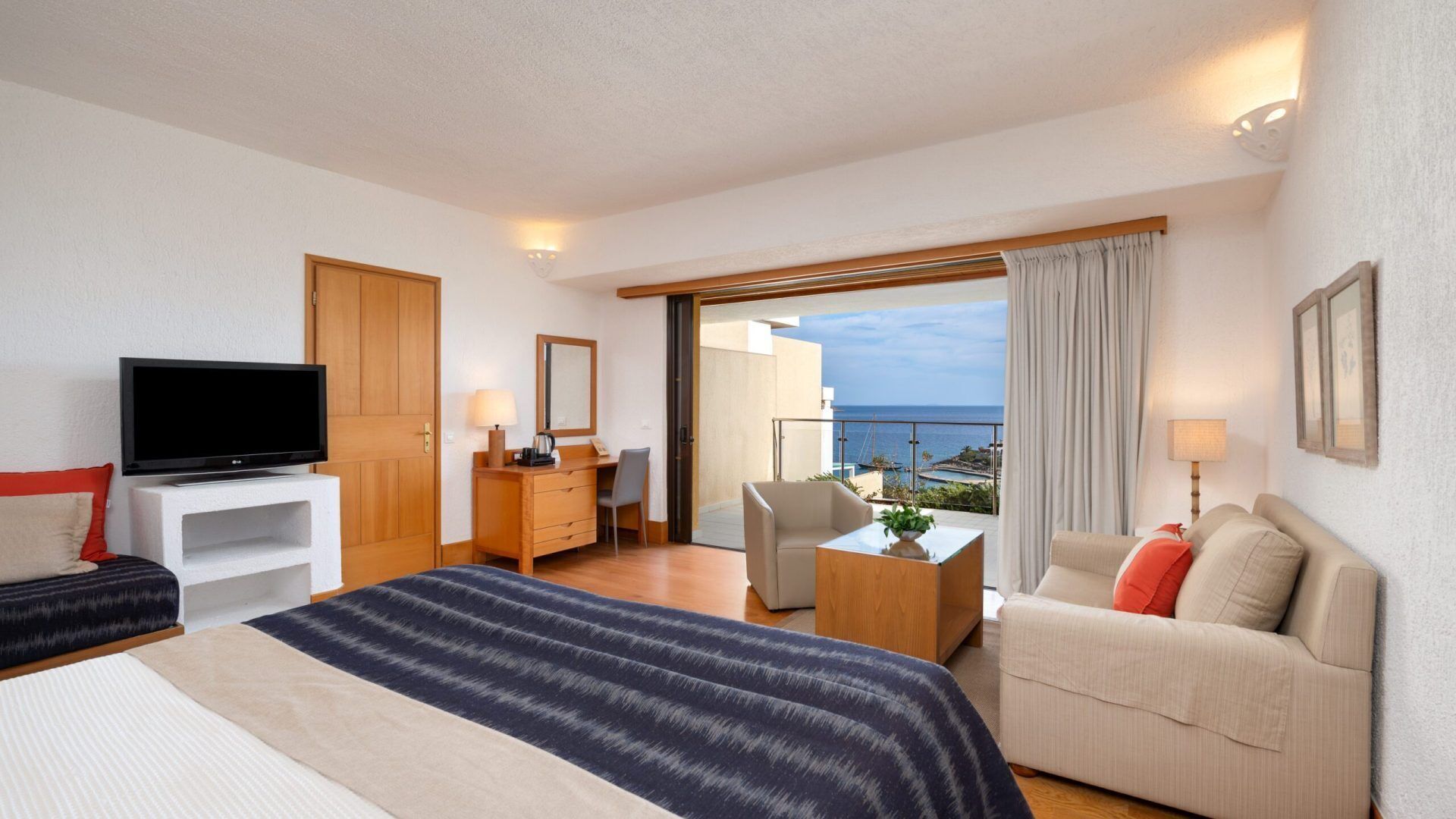 Ett hotellrum med en säng, en TV, ett skrivbord, en stol, en soffa och en balkong med utsikt över havet.