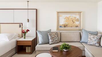 A modern hotel room with a bed, a nightstand with a vase of flowers, a sofa with patterned cushions, a coffee table with a book and a plant, and a framed artwork on the wall.