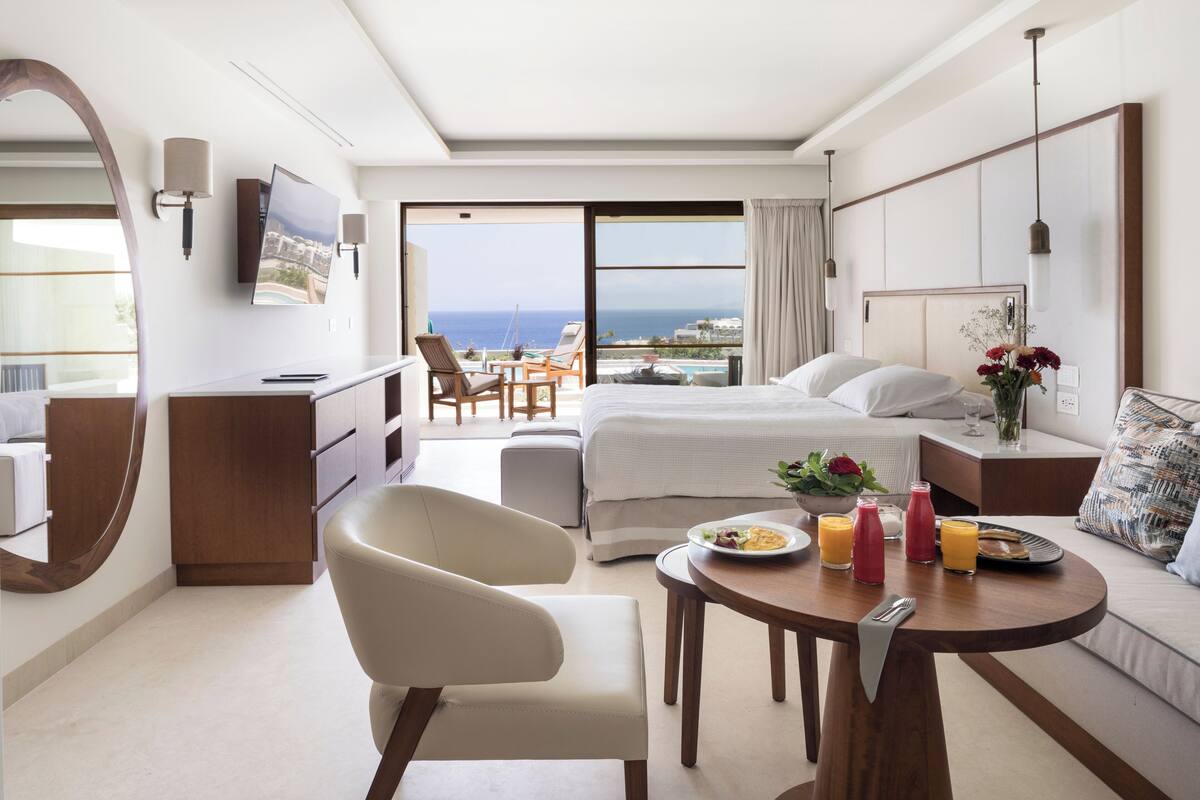 Et moderne hotelværelse med seng, skrivebord, stol, spisebord med morgenmad og havudsigt.