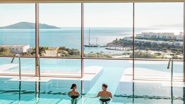 Piscina interna com grandes janelas que oferecem vista para o mar e edifícios distantes.