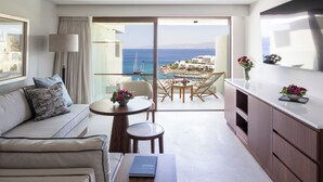 Uma sala de estar moderna com um sofá, uma mesinha de centro com um livro, uma varanda com vista para o mar e edifícios, e uma área de jantar com cadeiras e uma mesa.