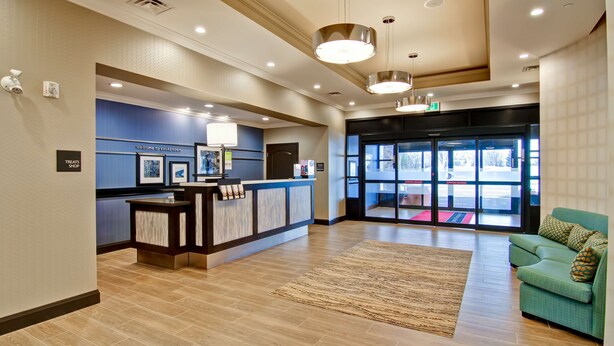 A reception area with a counter, a blue wall with framed pictures, a green sofa, and a large window with a view of the outside. - Hampton Inn & Suites by Hilton Saskatoon Airport (Saskatoon)