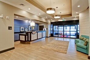 A reception area with a counter, a blue wall with framed pictures, a green sofa, and a large window with a view of the outside. - Hampton Inn & Suites by Hilton Saskatoon Airport (Saskatoon)