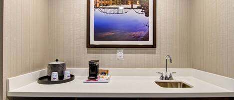 A modern bathroom with a white countertop, a sink, a coffee maker, and a framed picture of a bridge.