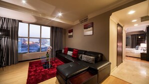 A modern living room with a black leather sofa, red and grey cushions, a glass coffee table, and a red rug. There are large windows with a city view, a door leading to a bedroom, and a wall with two artworks. - Best Western Plus Doha (Doha)