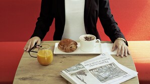 Une personne assise à une table, devant un croissant, une tasse de café et un journal.