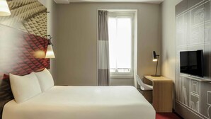 Une chambre d’hôtel avec un lit, une télévision, un bureau, une lampe et une fenêtre avec des rideaux.
