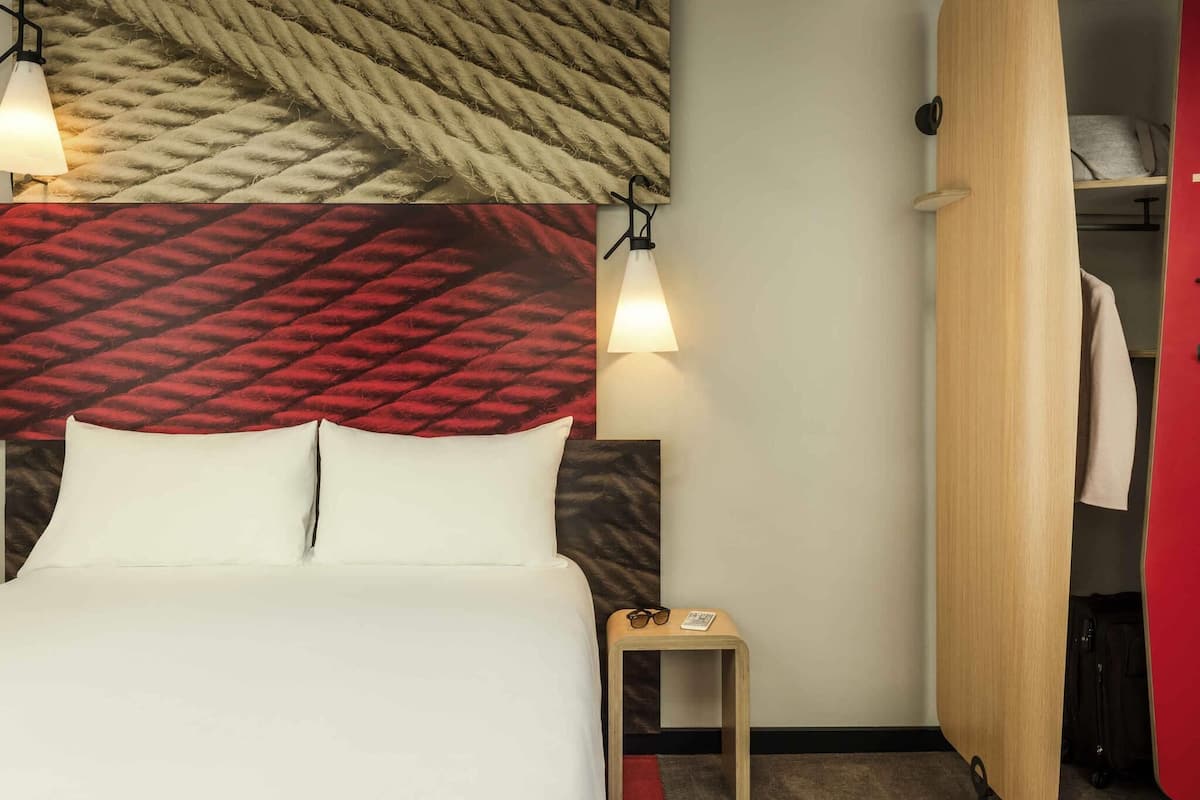 Hotelkamer met een bed, twee bedlampen, een klein tafeltje met glazen en een afstandsbediening, en een kast met een rode panelen.'