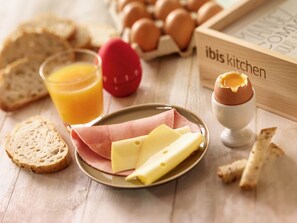 Une table en bois dressée pour le petit-déjeuner, avec du pain, des œufs, du fromage, du jambon et du jus de fruits.