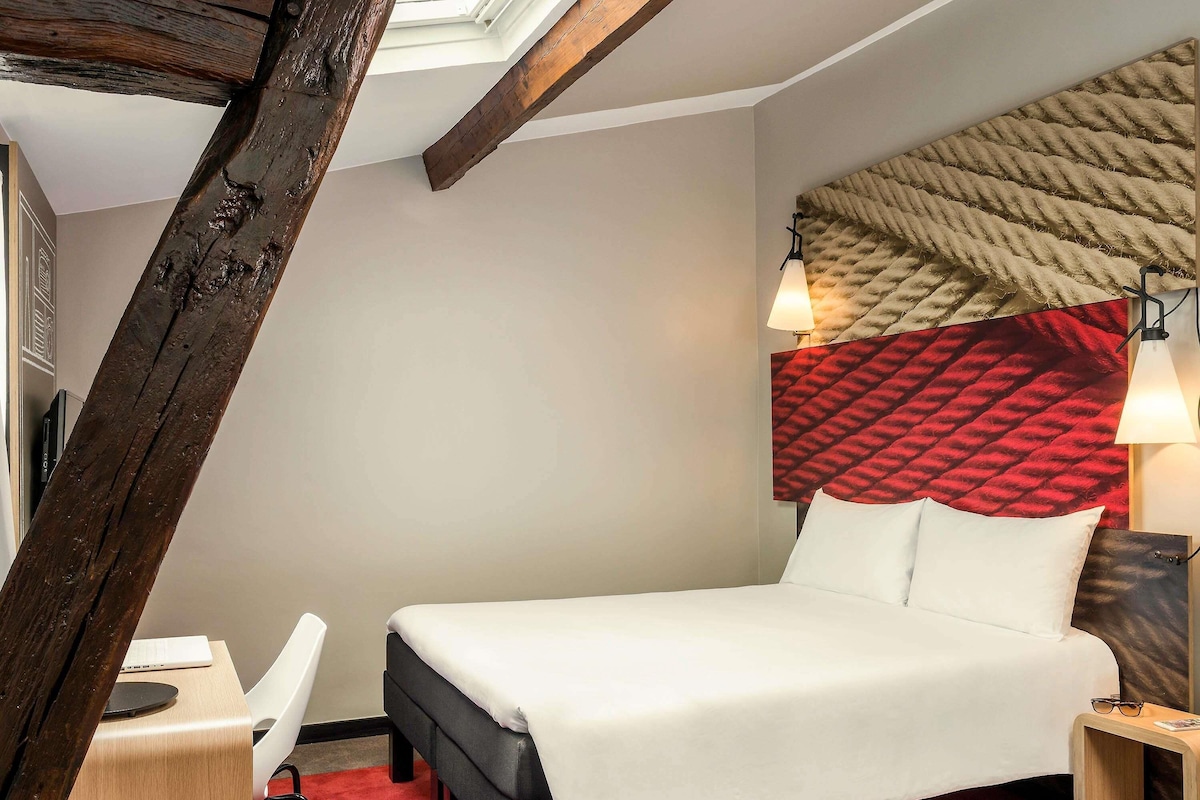 A hotel room with a bed, a desk, a chair, a lamp, and a headboard with a textured design.