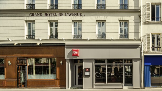 La façade du Grand Hôtel de Lavenue est ornée d’une enseigne bien visible indiquant « Grand Hôtel de Lavenue », placée au-dessus de l’entrée, ainsi que du logo « ibis » sur la droite, et est ponctuée de fenêtres et de balcons.