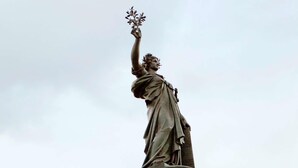 Une statue représentant une figure tenant une branche ornée de feuilles, posée sur un piédestal décoré de sculptures complexes.