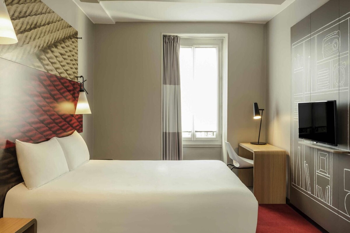 Een hotelkamer met een bed, een televisie, een bureau, een lamp en een raam met gordijnen.