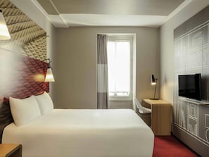 Ett hotellrum med en säng, en tv, ett skrivbord, en lampa och ett fönster med gardiner.