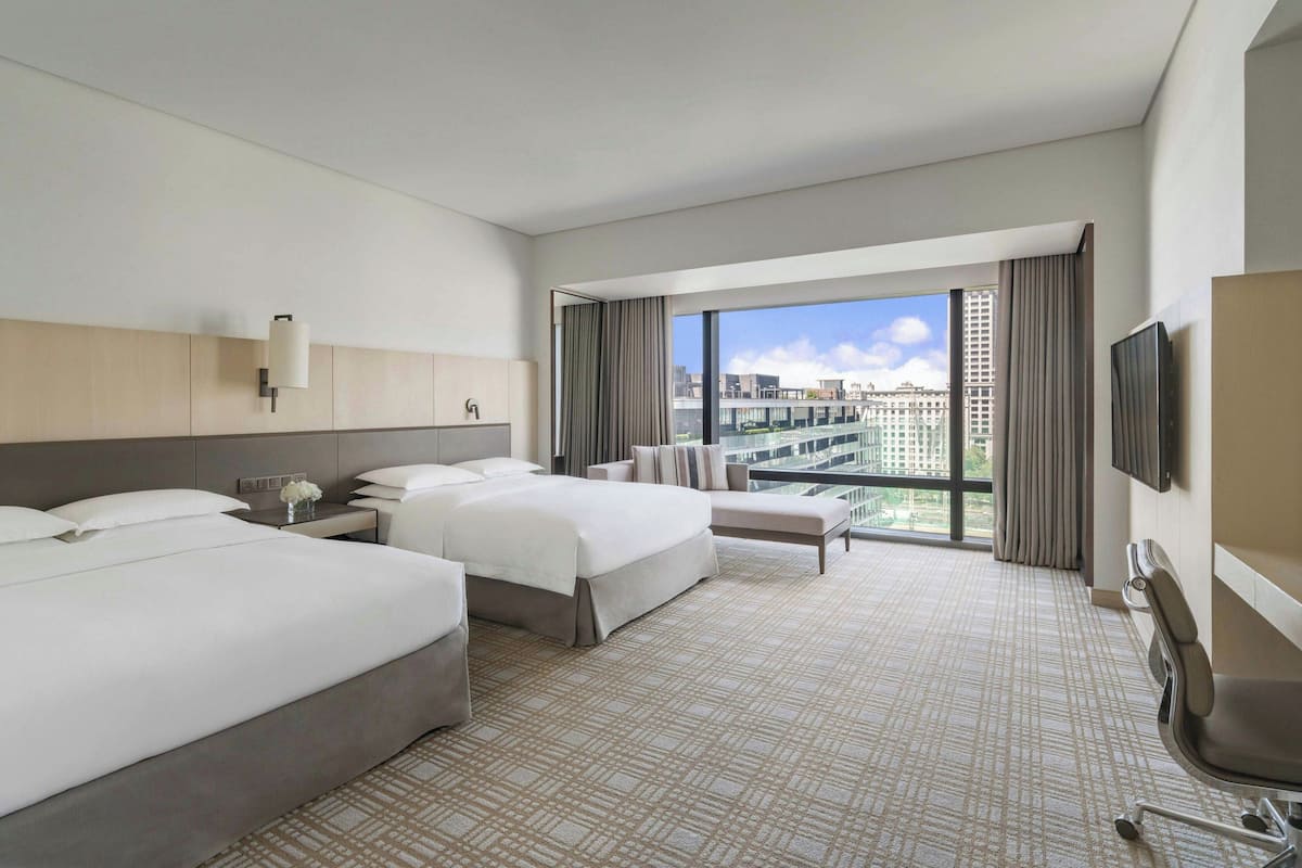 A hotel room with two beds, a sofa, a TV, and a large window with a city view.