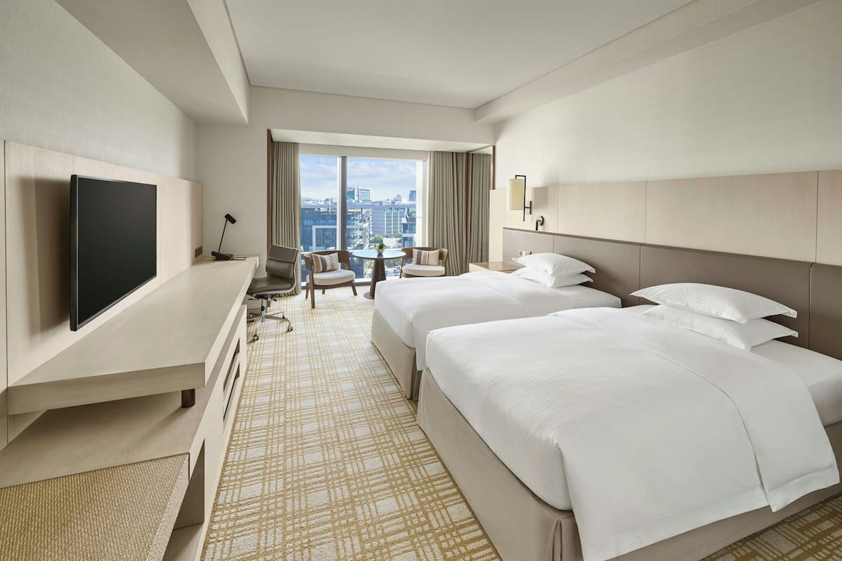 A hotel room with two beds, a TV, a desk, and a city view.