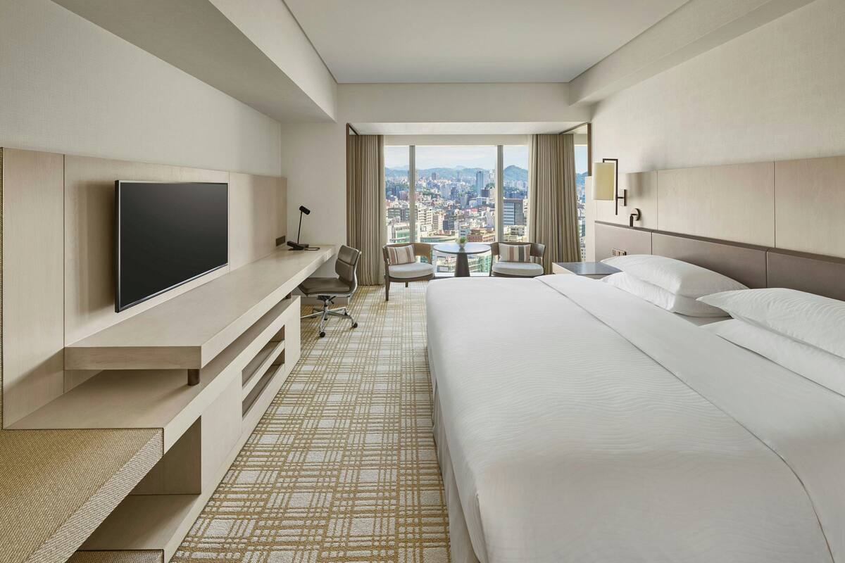 A hotel room with a bed, TV, seating area, and a city view.