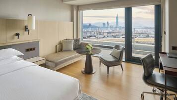 A modern hotel room with a large bed, a seating area with a table, and a view of a cityscape through large windows.