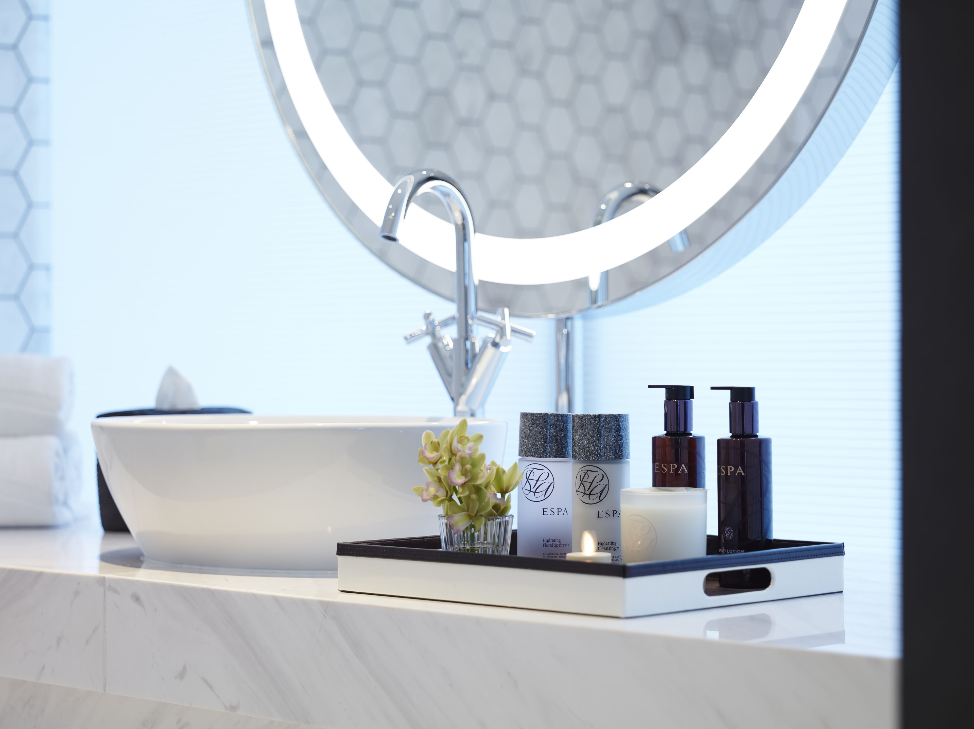 Un baño moderno con un lavamanos grande de color blanco, un espejo redondo y un jarrón pequeño con flores.