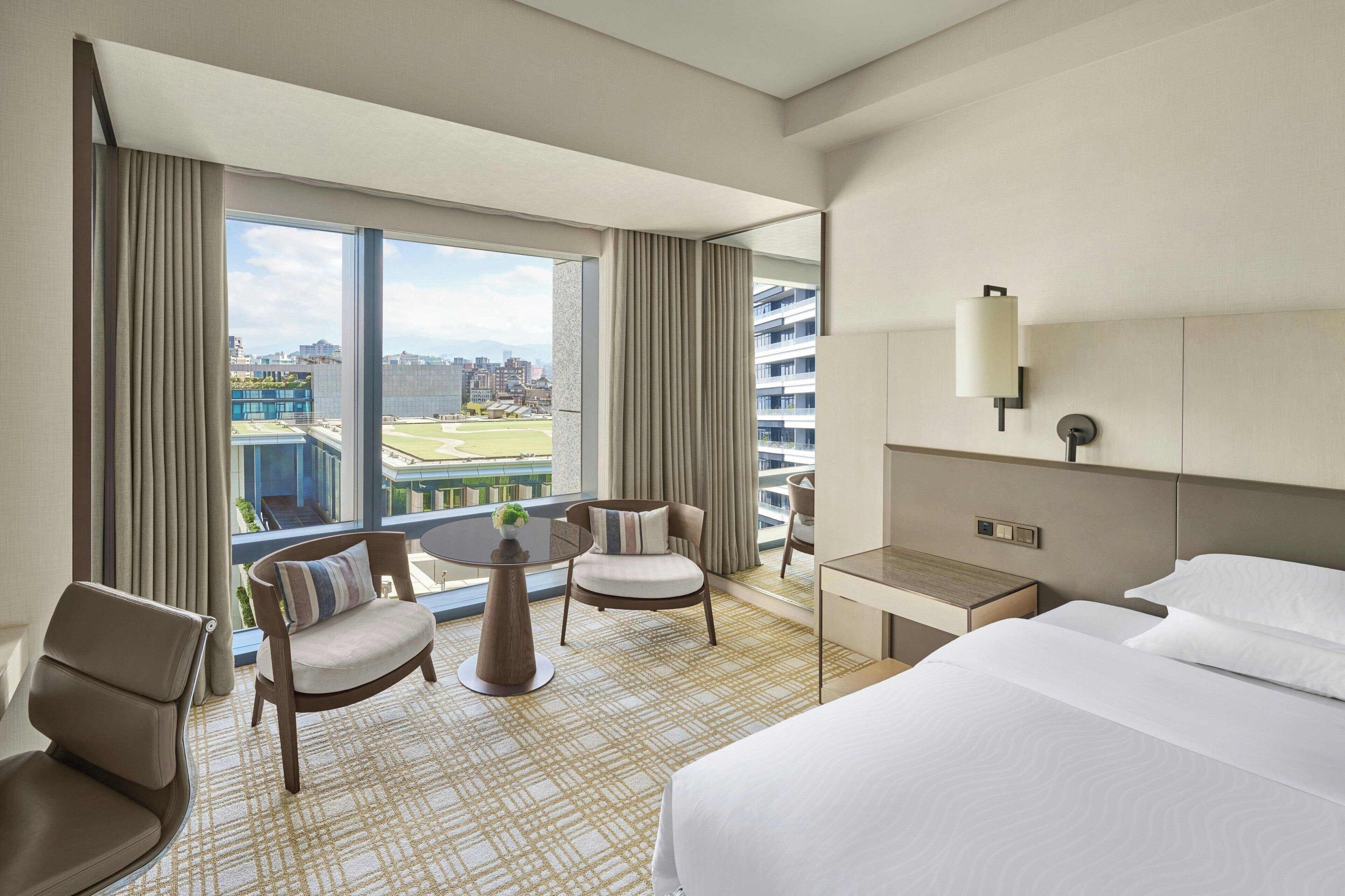 A hotel room with a bed, a desk, a chair, a small table, and a view of a cityscape.