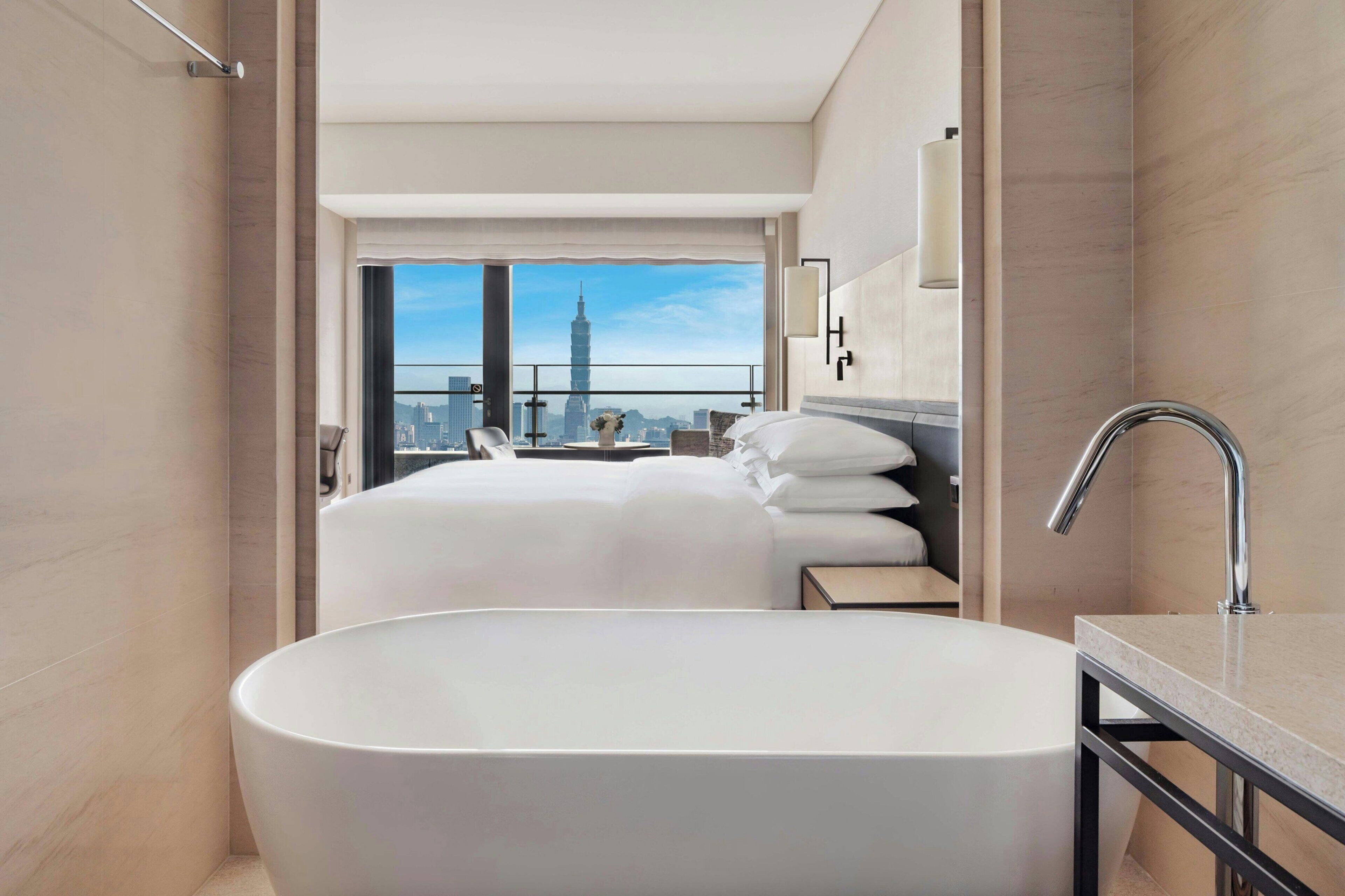 A modern hotel room with a large bathtub, a bed with white linens, a view of a city skyline, and a bathroom with a large mirror and chrome fixtures.