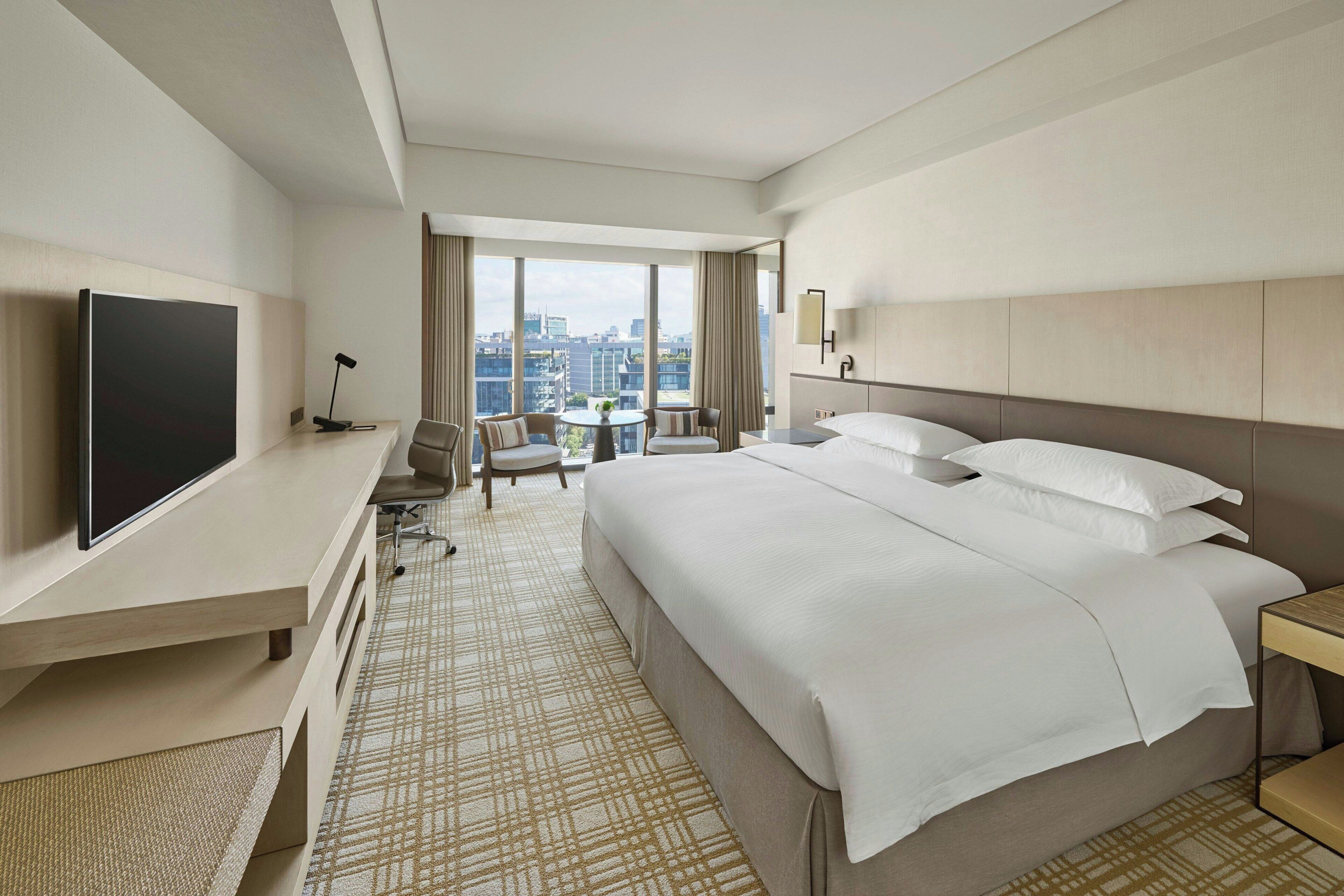 A hotel room with a large bed, a desk with a flat-screen TV, a chair, a small table with a plant, and a view of the city through large windows.