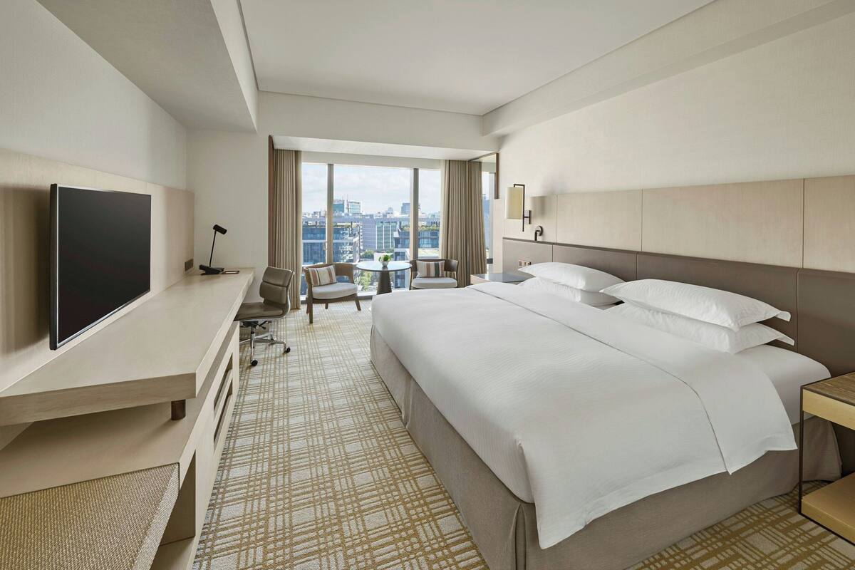 A hotel room with a large bed, a desk with a flat-screen TV, a chair, a small table with a plant, and a view of the city through large windows.