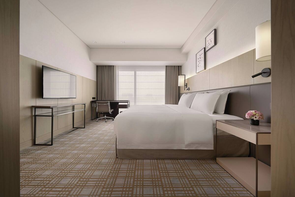 A hotel room with a large bed, a desk with a chair, a TV mounted on the wall, and a window with curtains.