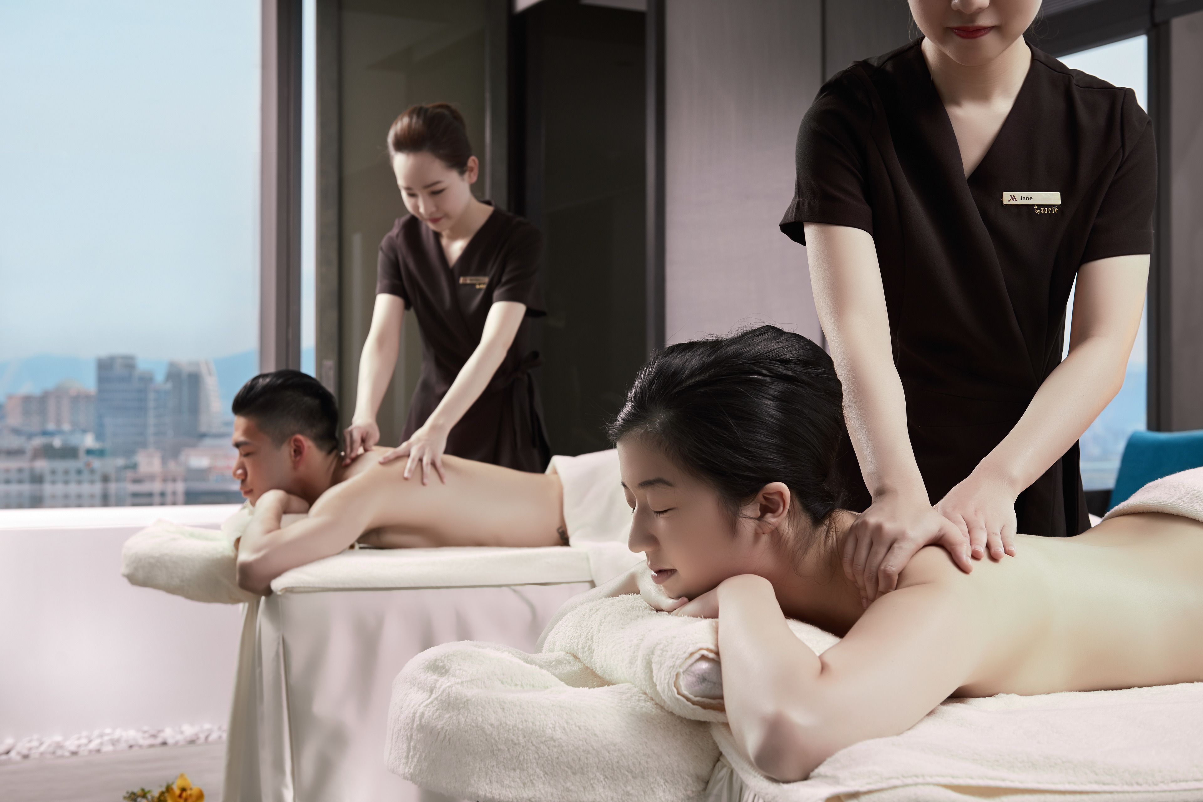 A massage therapy room with two clients receiving back massages from therapists, a city view through large windows, and a modern interior design.