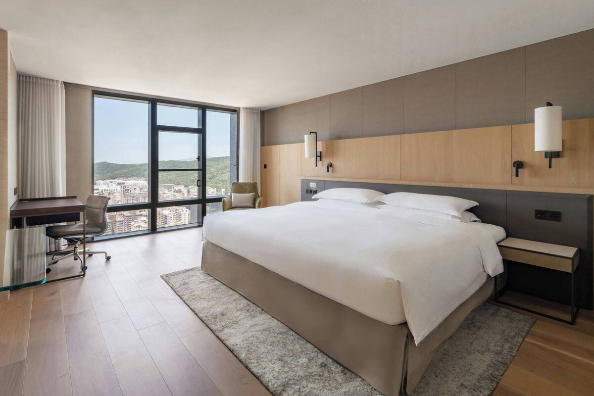 A modern hotel room with a large bed, a desk, and a view of the city.