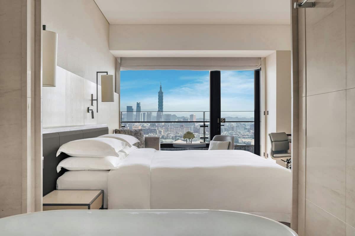 A modern hotel room with a large bed, a small table, a chair, and a view of a cityscape.