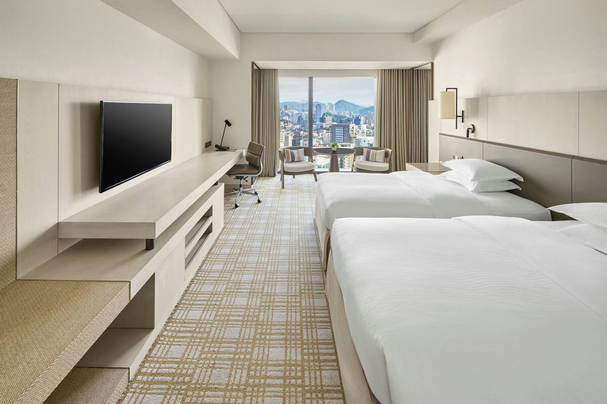 A hotel room with a flat-screen TV, two beds, a desk, and a chair.