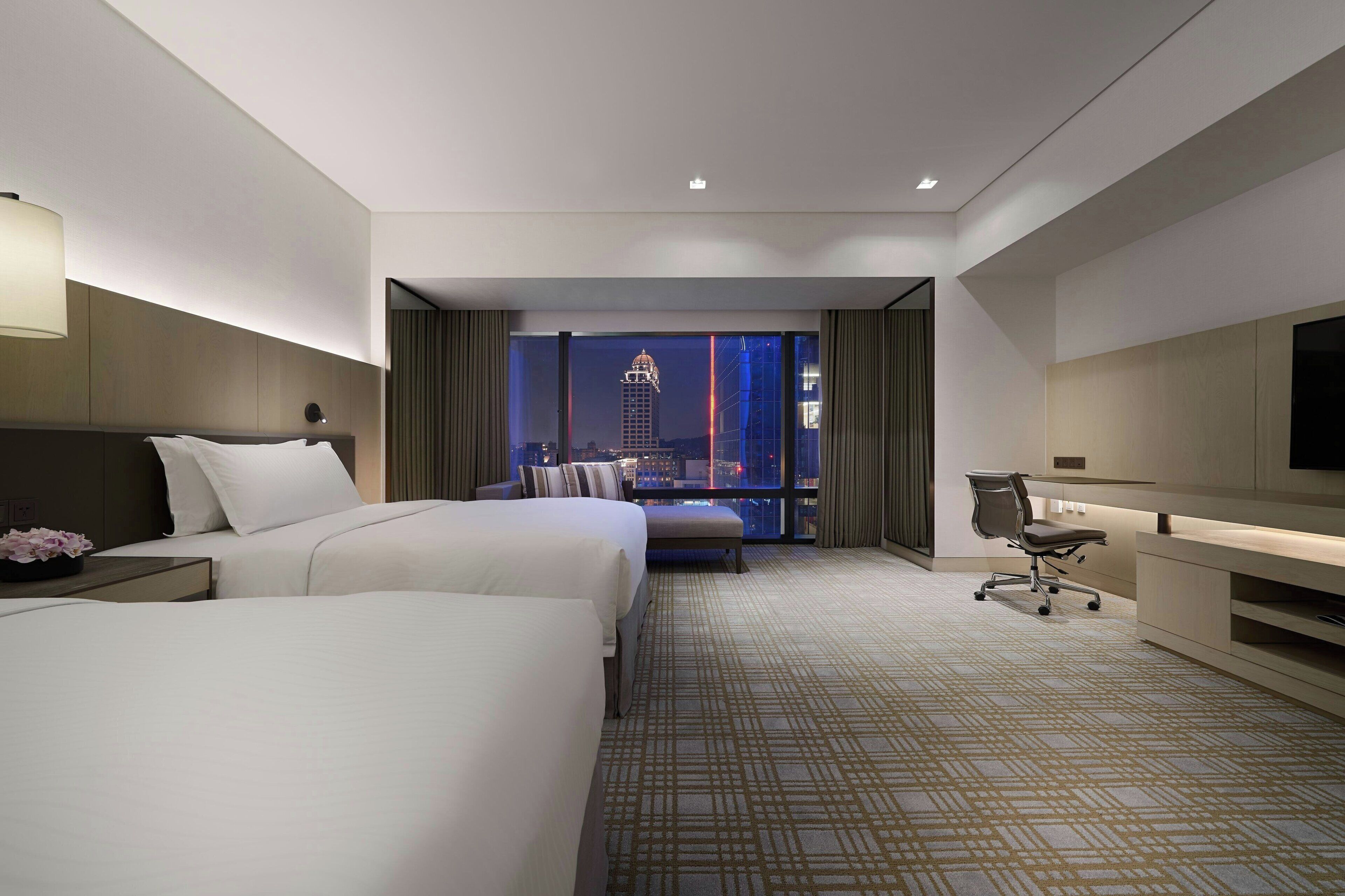 A hotel room with two beds, a desk, a chair, and a large window with a city view.