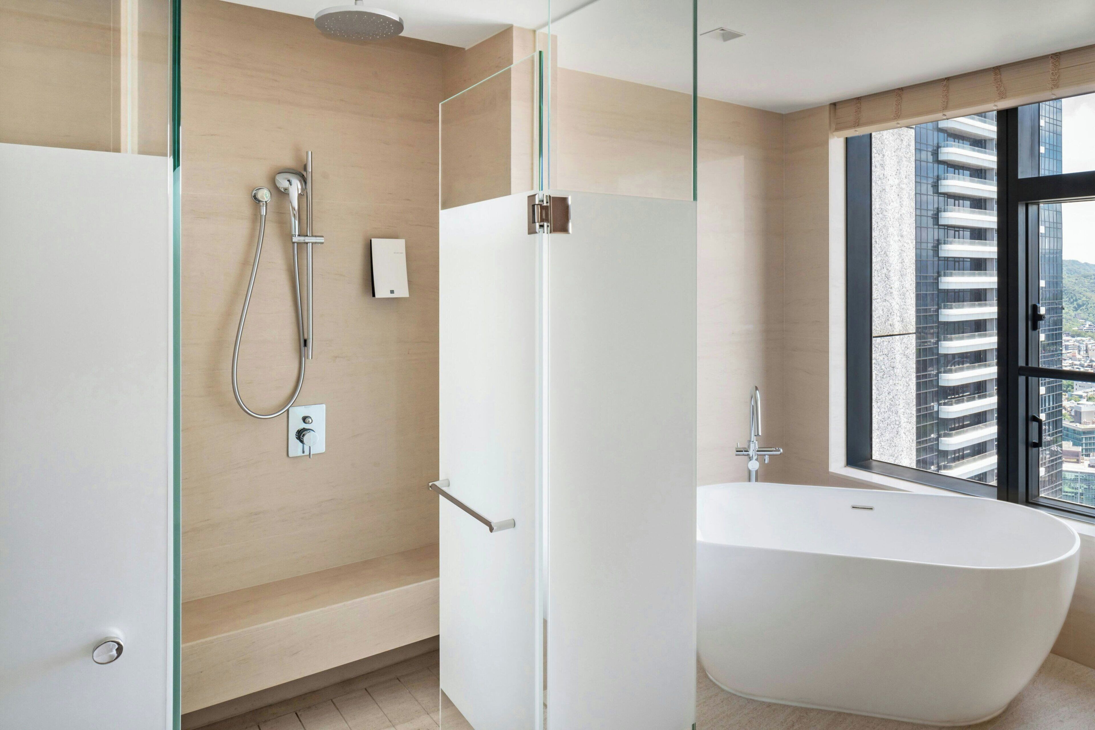 A modern bathroom with a glass-enclosed shower, a freestanding bathtub, and a large window offering a city view.
