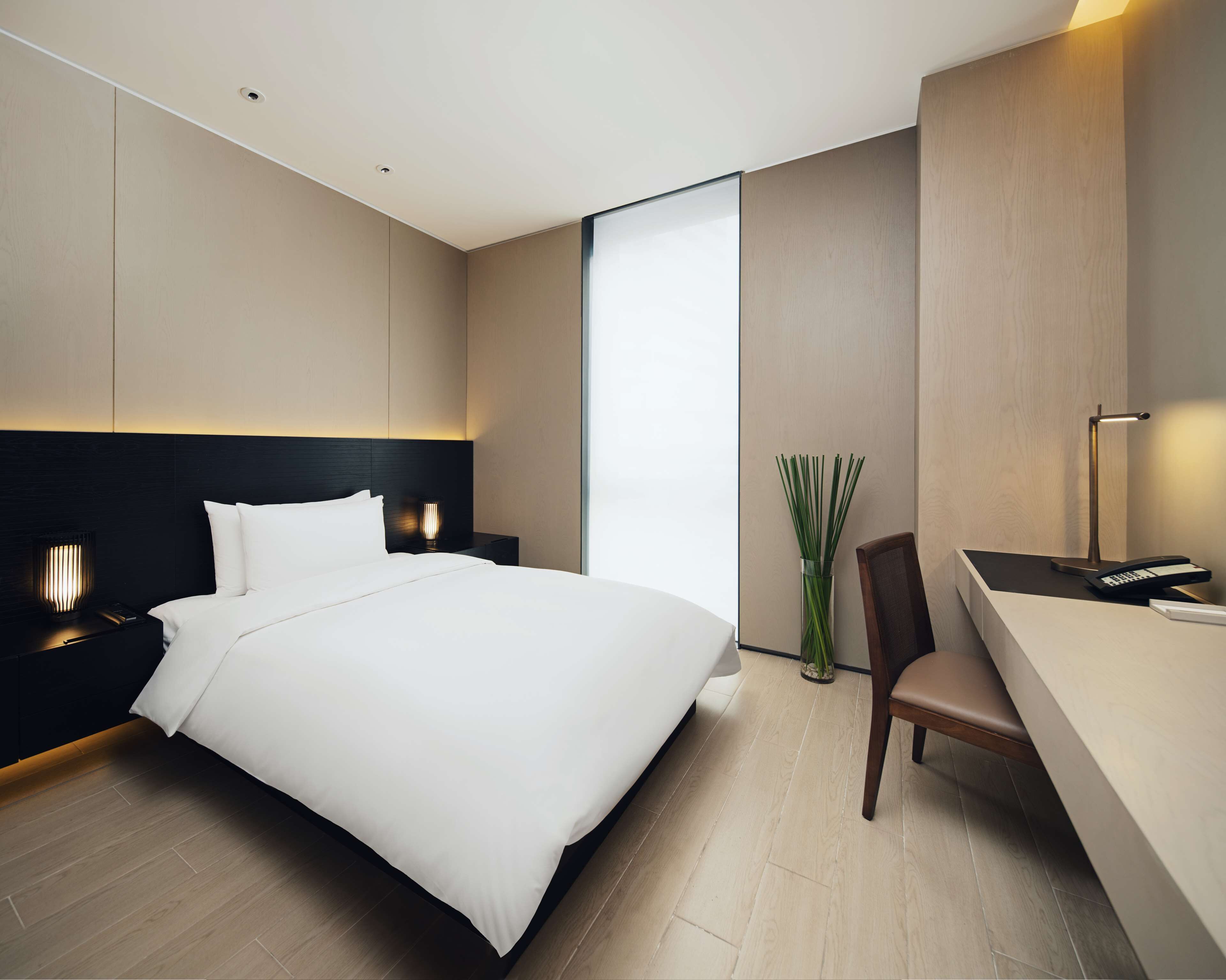 A modern hotel room with a large bed, a desk with a chair, a vase with green plants, and a nightstand with a lamp.