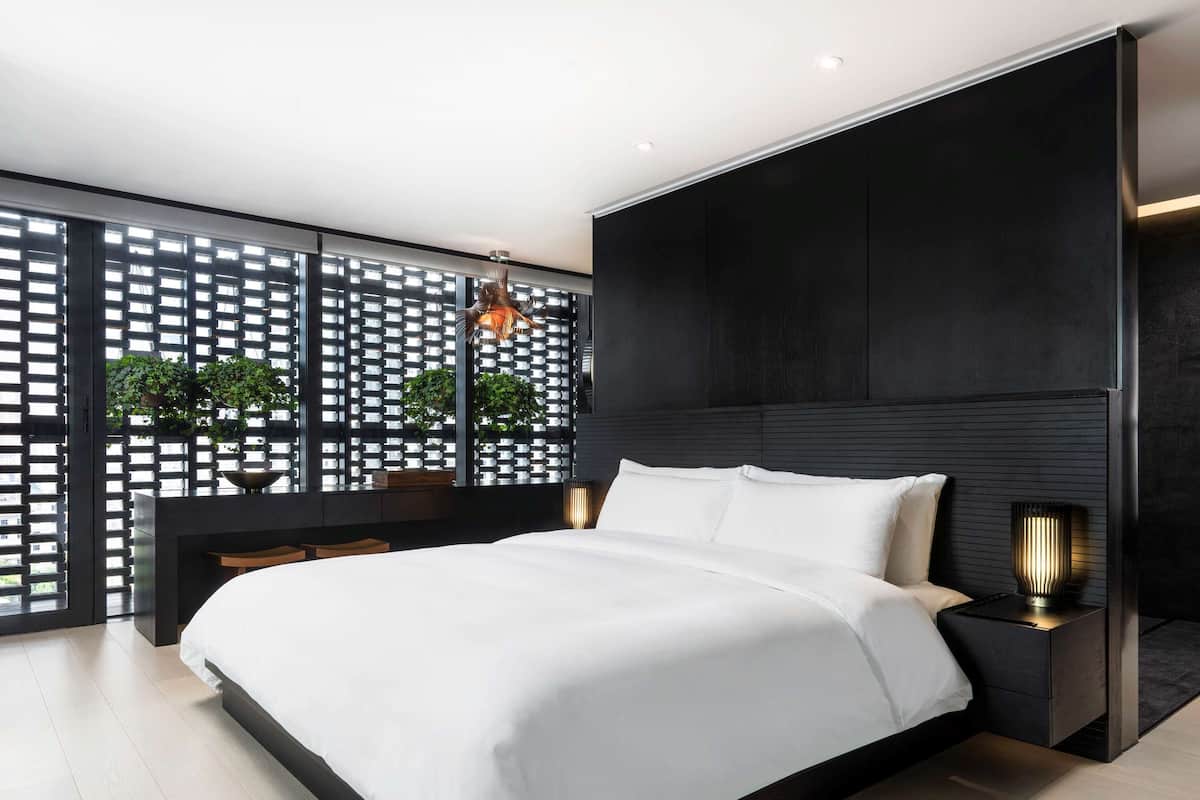 A modern bedroom with a large bed, a dark headboard, a nightstand with a lamp, and a view through a lattice window with plants.