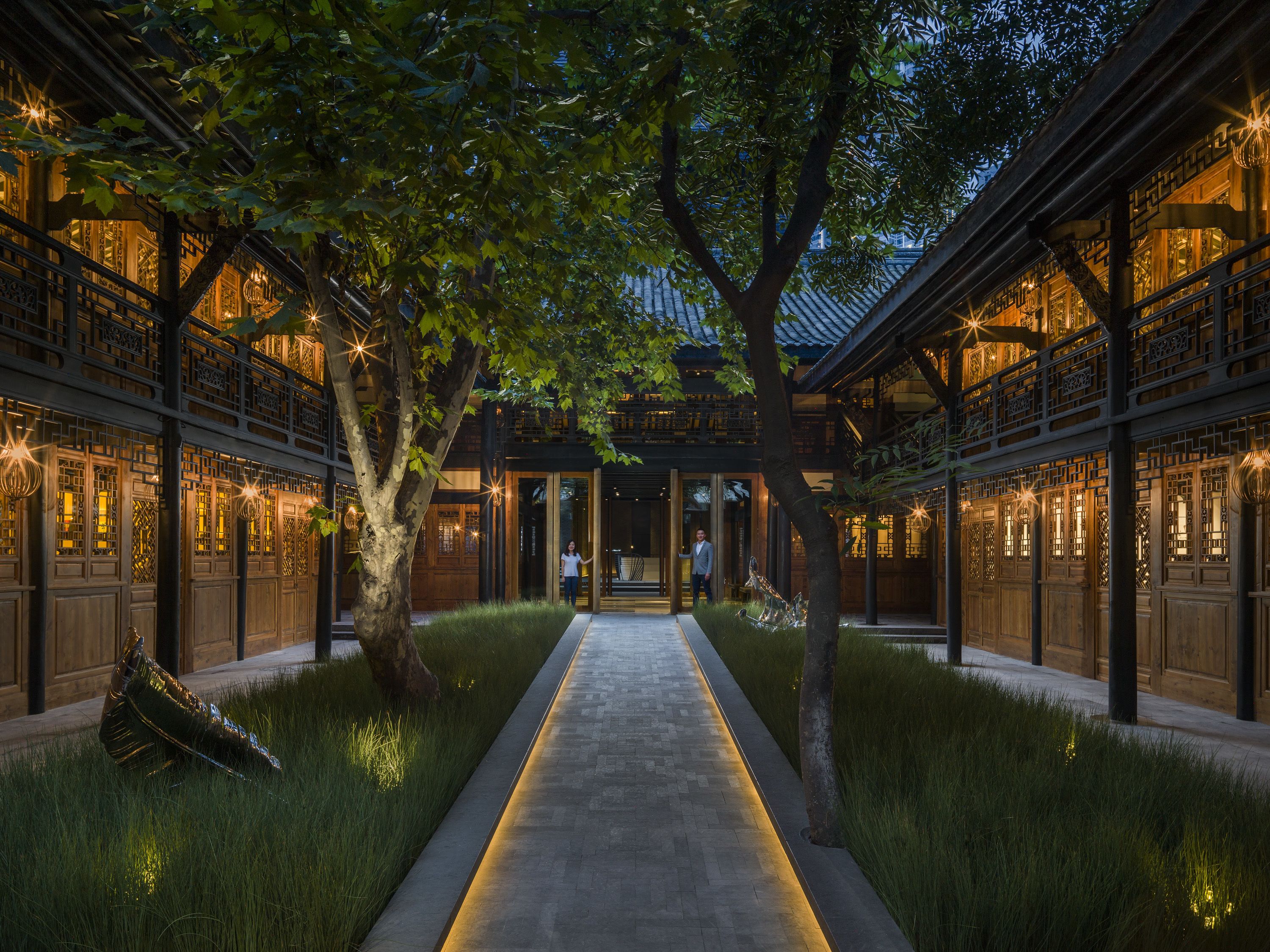 A traditional courtyard with a central pathway, wooden buildings with intricate lattice windows, and a tree in the middle.