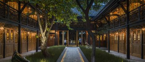 A traditional courtyard with a central pathway, wooden buildings with intricate lattice windows, and a tree in the middle.