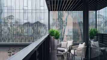 Un balcón moderno con mobiliario de mimbre, una planta en maceta y vista panorámica de la ciudad a través de grandes ventanales.