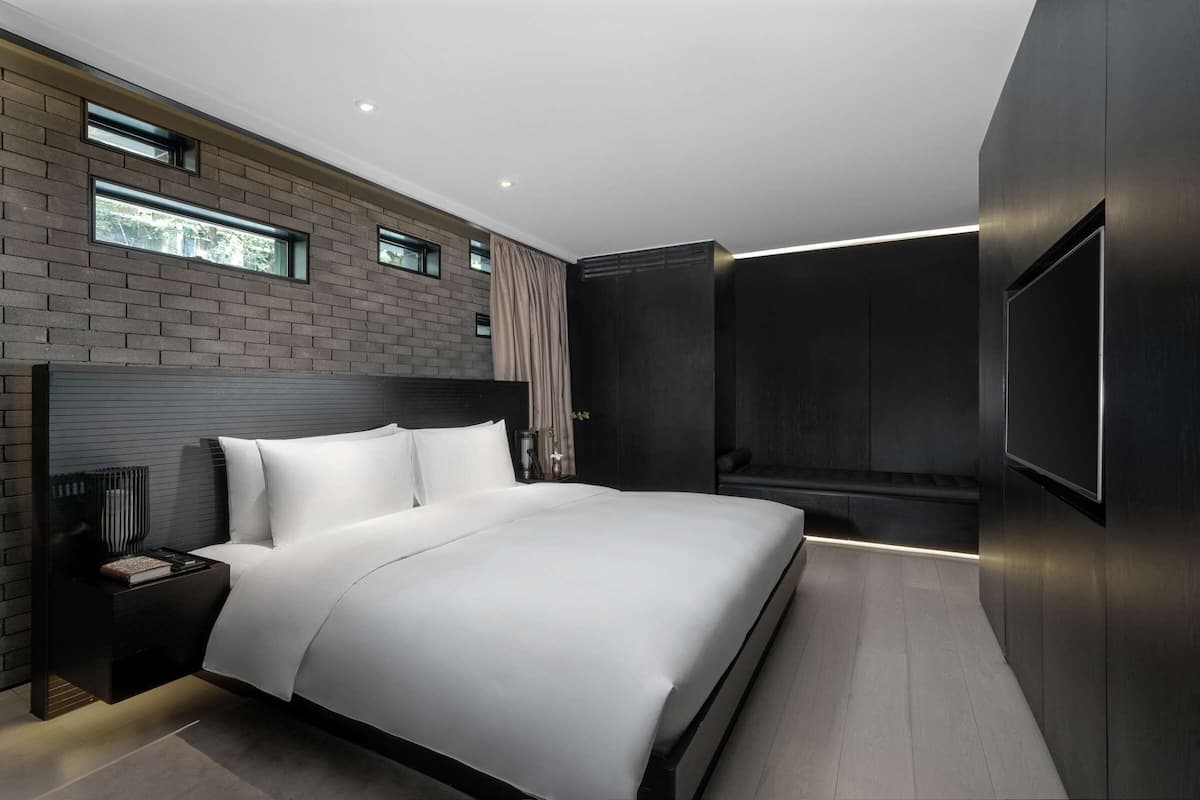 A modern bedroom with a large bed, a nightstand, a built-in TV, and a window with a view of greenery.