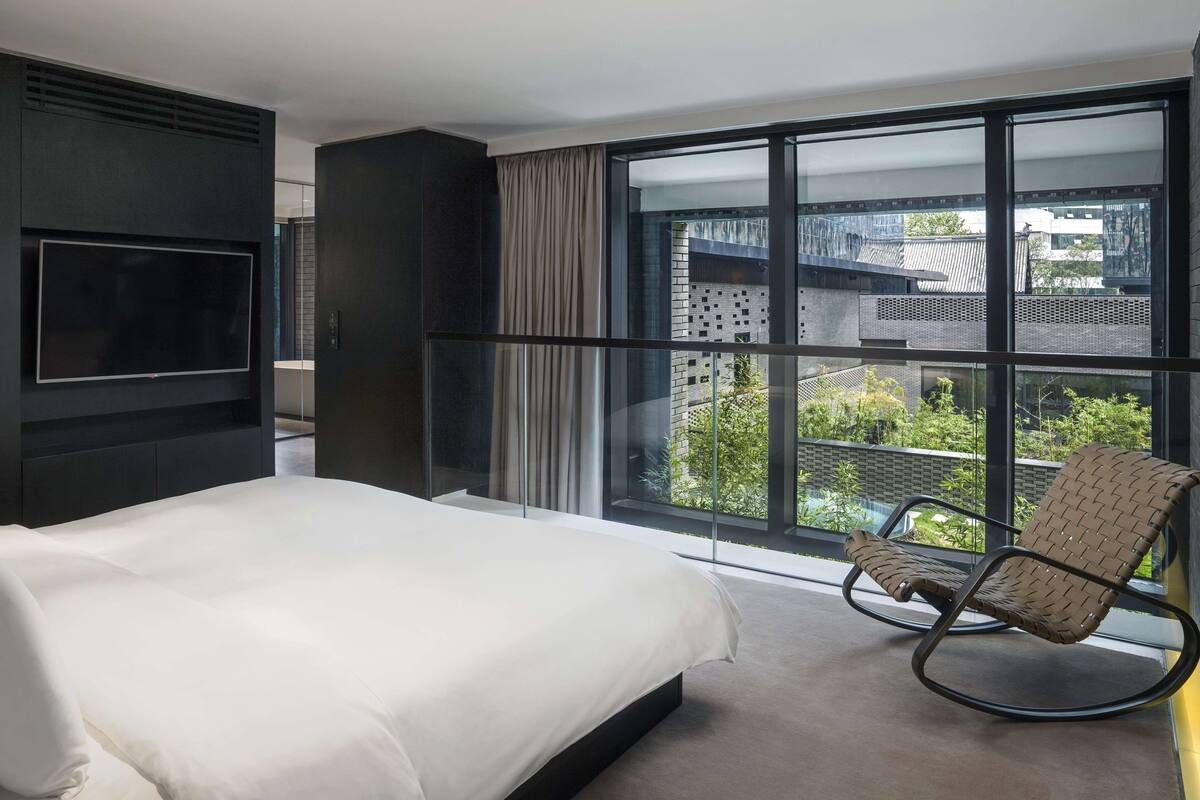 A modern hotel room with a large bed, a TV mounted on the wall, a balcony with a chair, and a view of the cityscape.