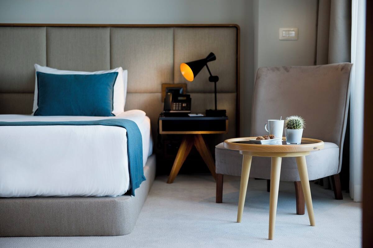 Une chambre d’hôtel avec un lit, une table de chevet, une lampe, une chaise et une petite table sur laquelle se trouvent une tasse et une plante.