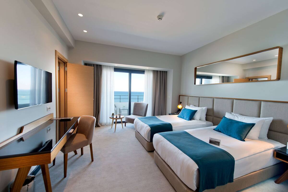 Une chambre d’hôtel avec deux lits, un bureau, une chaise, une télévision et une grande fenêtre avec des rideaux.