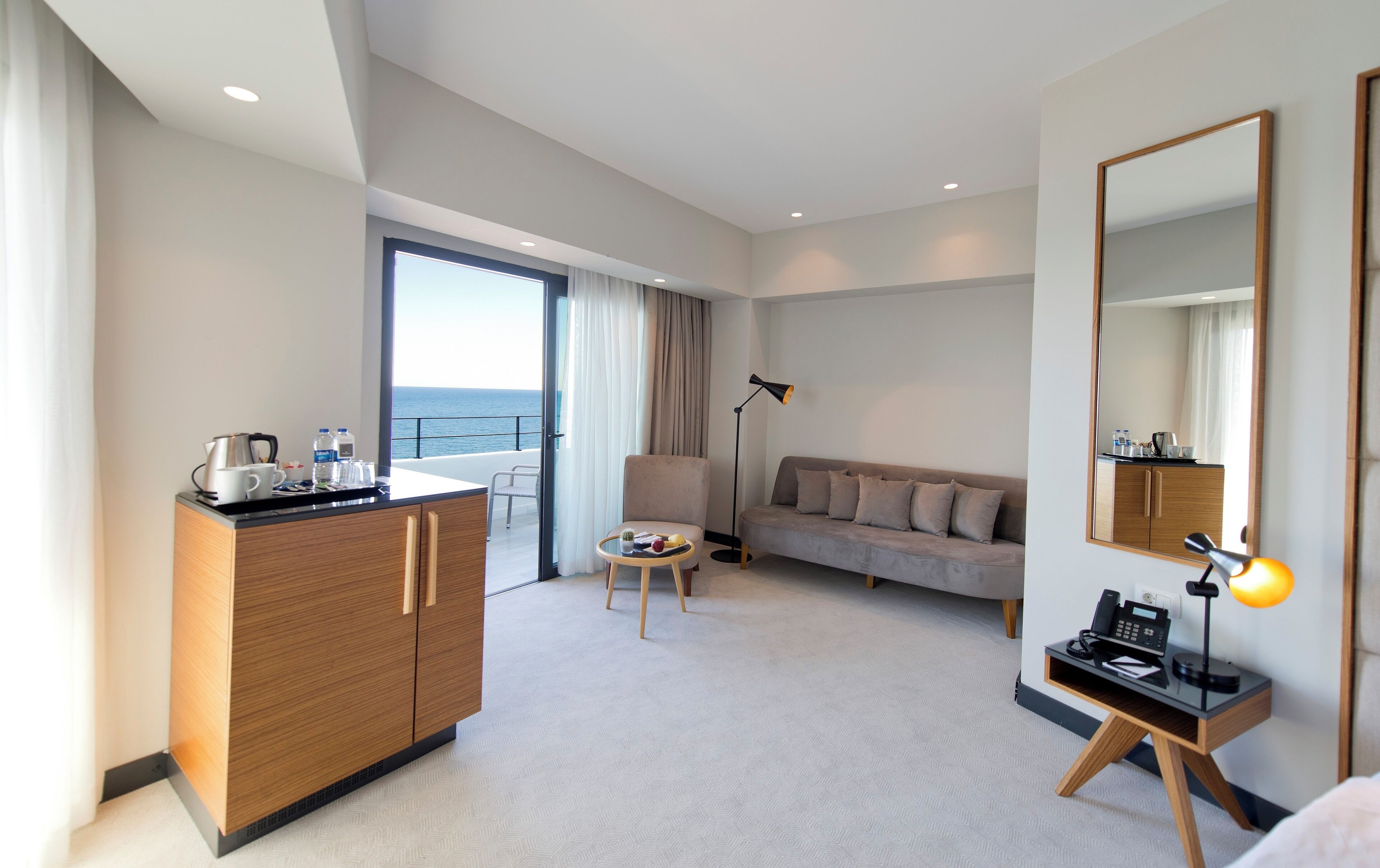 Una habitación de hotel moderna con un armario de madera, un sofá, una mesita con una bandeja, una lámpara de pie y un espejo grande.